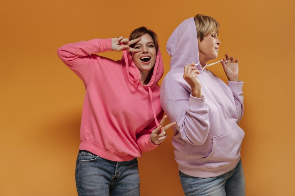 Cheerful young girl in pink sweatshirt showing peace signs, winking and posing with modern woman in