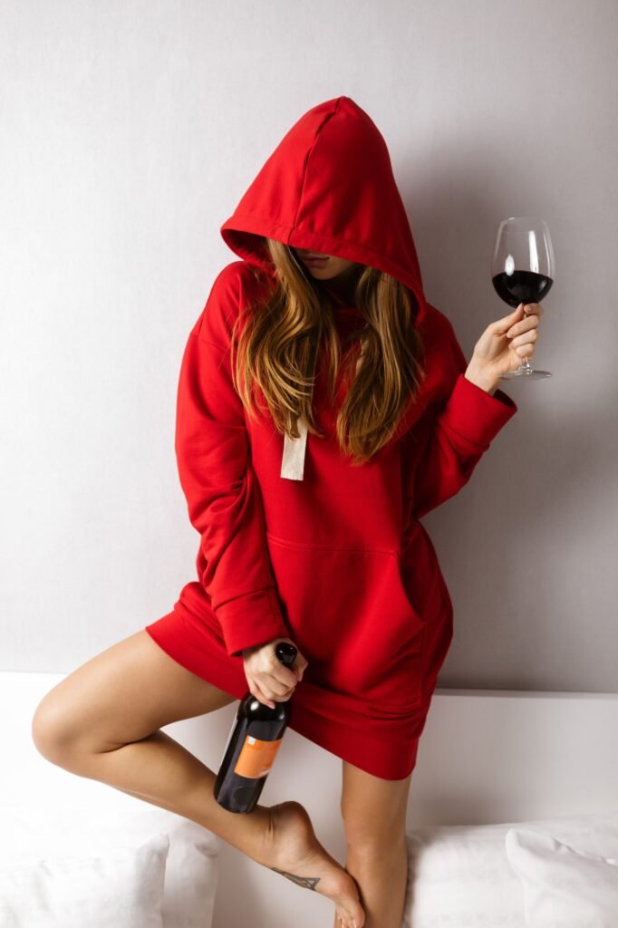Young lady in red hoodie standing in bed and holding bottle and glass of red wine in hands